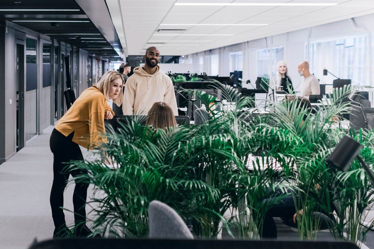 A modern office space with several people working and interacting. The office has a long corridor with workstations on both sides, separated by green plants. The ceiling has white panels and the floor is carpeted in gray. There are overhead lights providing illumination.